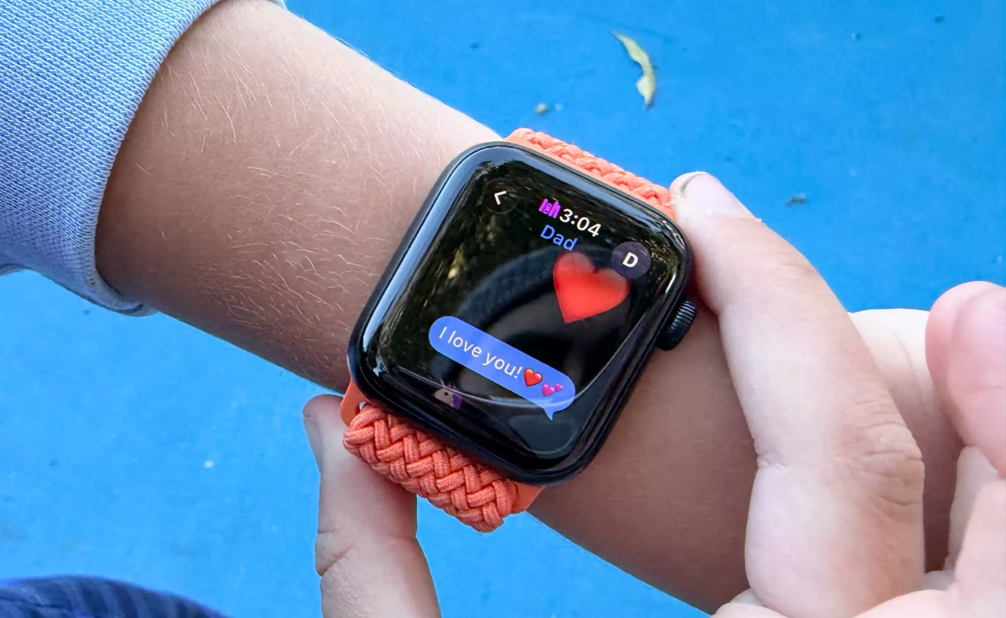 A child's wrist wearing a connected watch, messaging I love you to Dad on a blue court